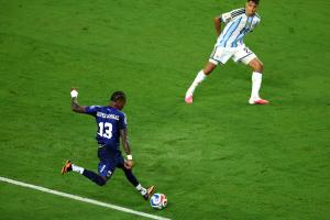 a soccer player in a navy jersey winds up to take a shot as a defender braces to block