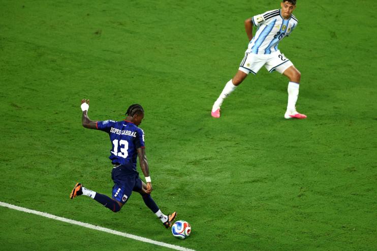 a soccer player in a navy jersey winds up to take a shot as a defender braces to block