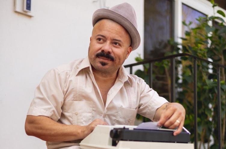 photo of a man in a button-down shirt and fedora typing on a typewriter on a stoop