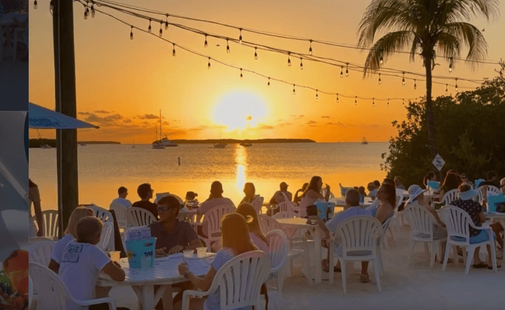 a waterfront outdoor restaurant on the sand with the sunset in the background