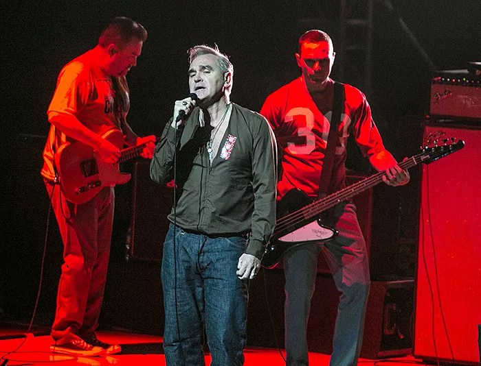 photo of singer Morrissey singing onstage with two musicians behind him bathed in red light