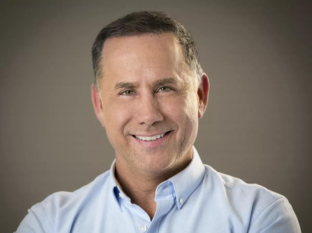 a whiteman in a light blue button-down smiles at the camera in front of a grey background