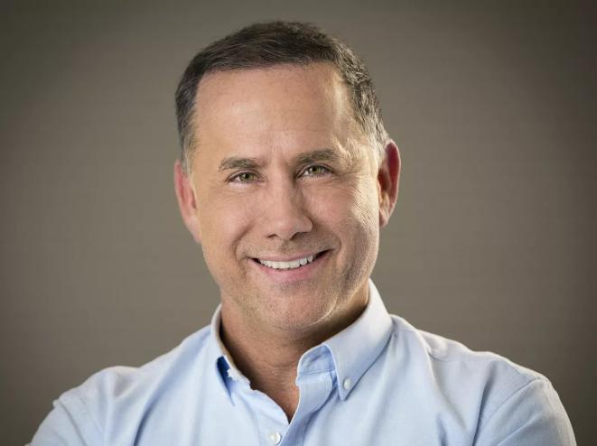 a whiteman in a light blue button-down smiles at the camera in front of a grey background