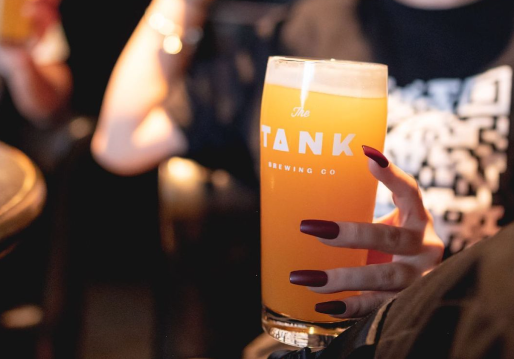 close-up photo of a hand with long, red nails holding a pink of beer with a logo on the glass reading "Tank"