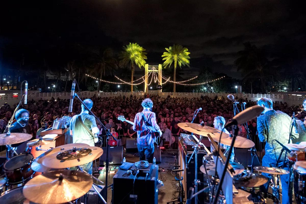 photo of a band performing onstage in front of an outdoor audience at night from the view of the drummer