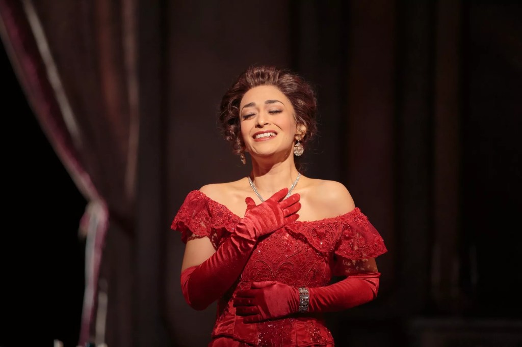 photo of an opera singer in a red dress and red gloves singing onstage with her hand to her chest