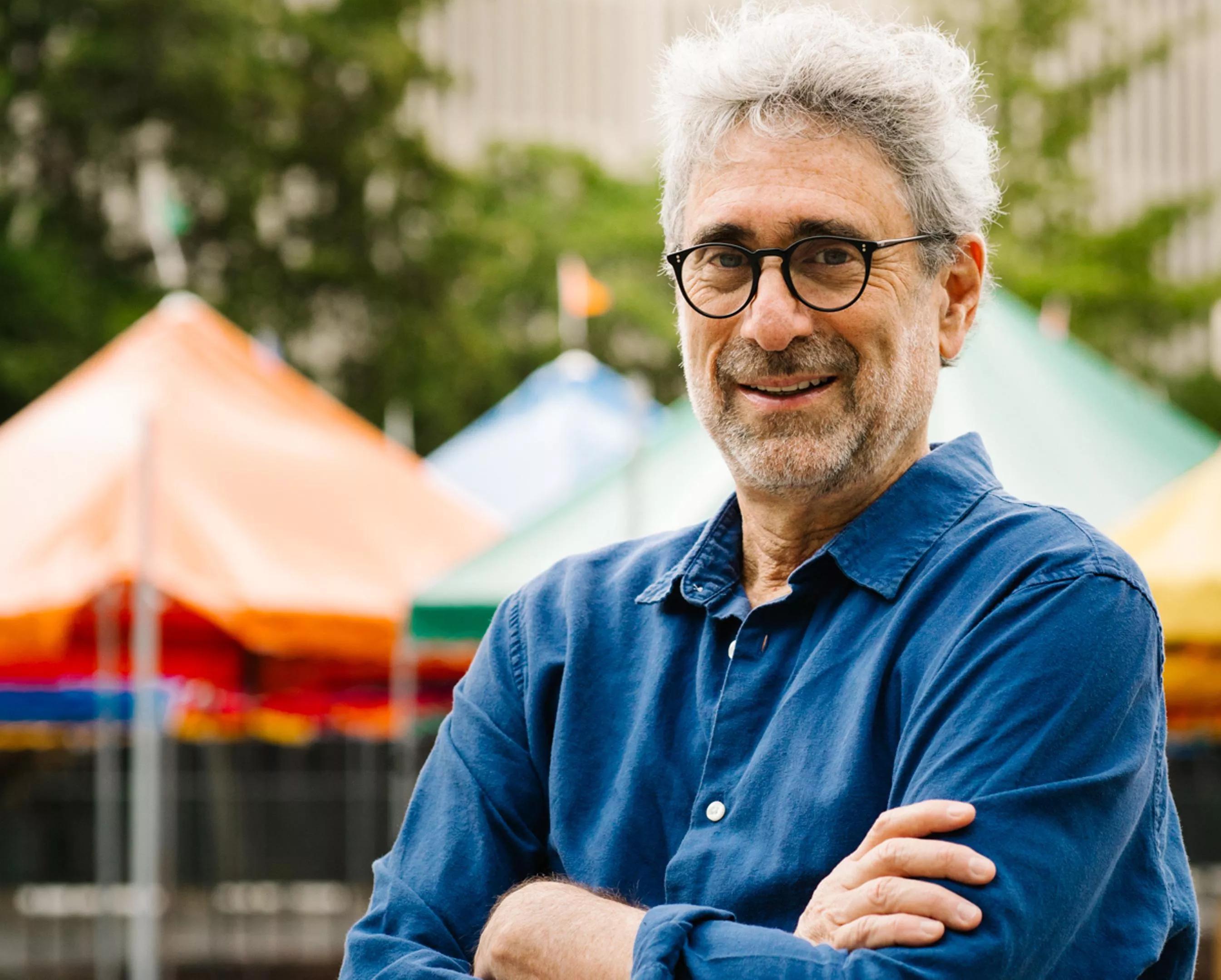 Color photo of Mitchell Kaplan, founder of Books & Books, an independent bookstore chain based in Coral Gables (Miami), Florida. In this horizontal image, the bespectacled bookseller stands with arms crossed, in front of tents set up in downtown Miami for the annual Miami Book Fair, an event Kaplan cofounded in 1984.