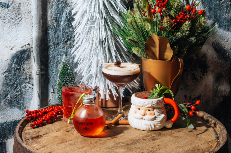 A group shot of holiday cocktails made with Patron. The drinks sit on a wooden stool surrounded by holiday decor.