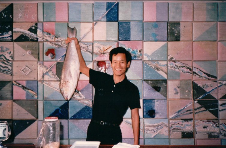 vintage photo of a smiling man dressed in black, standing at a counter against a tiled background and holding up a whole fish in his right hand