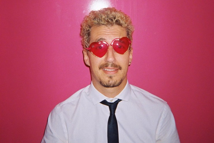 Josch wears heart-shaped sunglasses standing against a bright pink background
