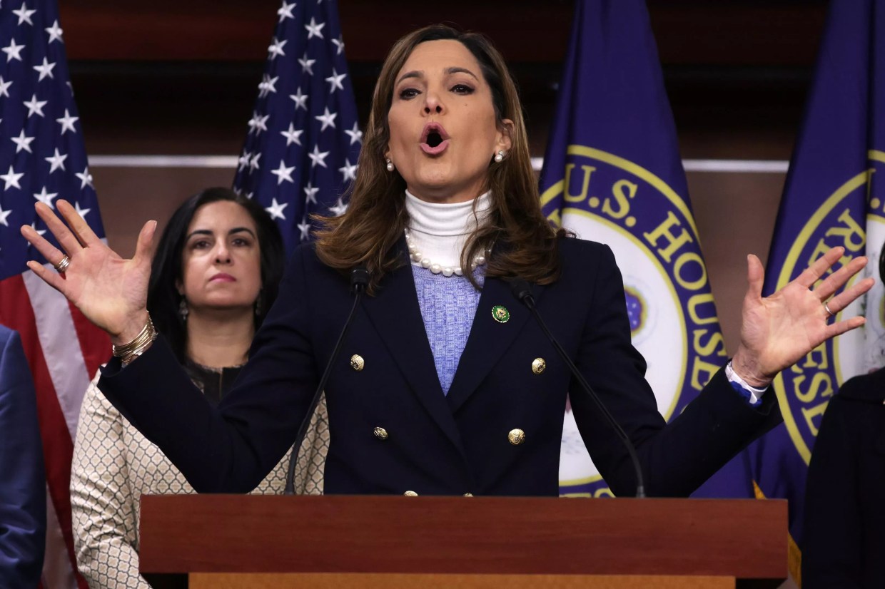 A woman in a navy blazer speaks from a wooden podium with American flags behind her