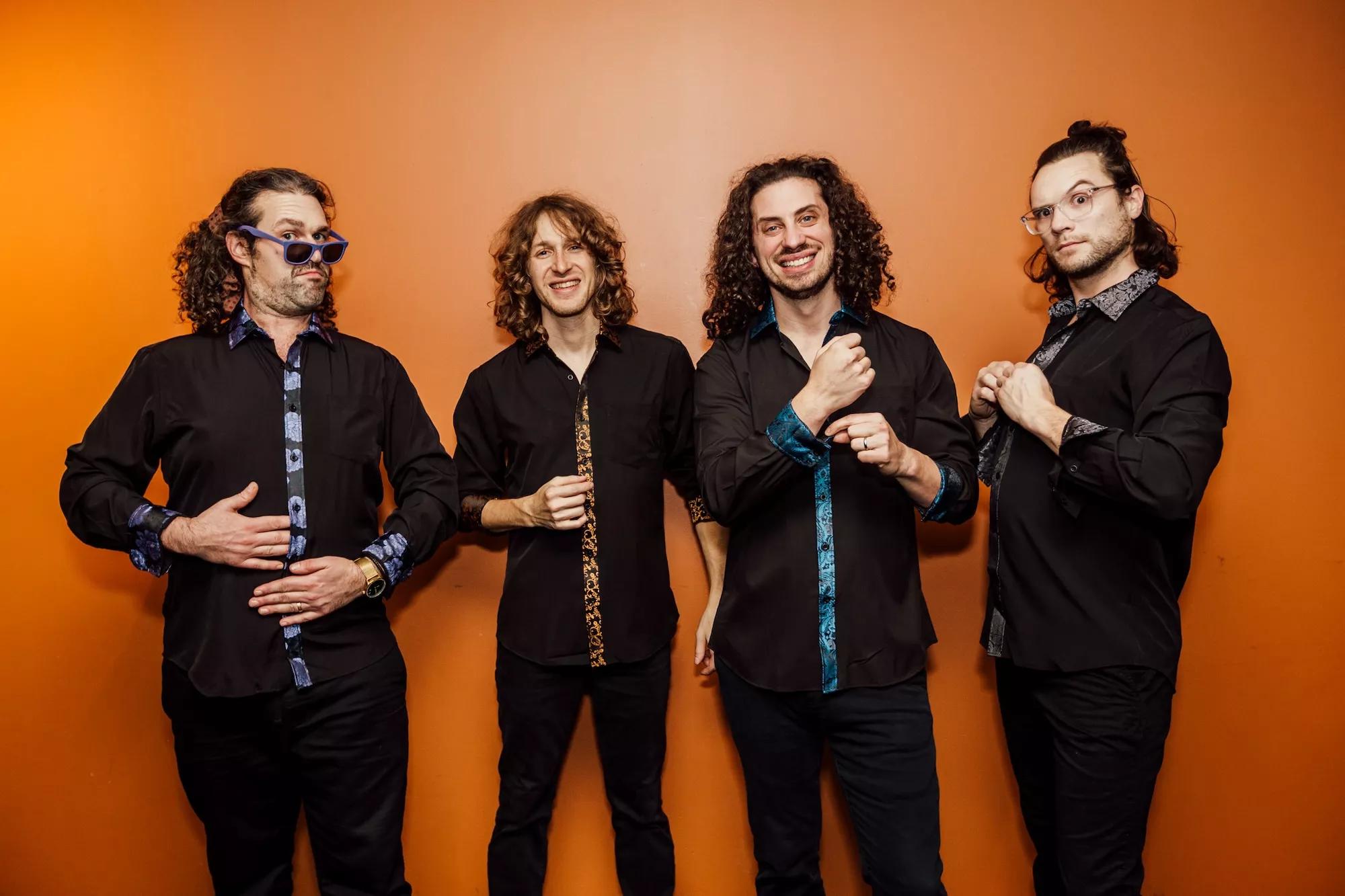 The four members of Pigeons Playing Ping Pong against a bright orange wall