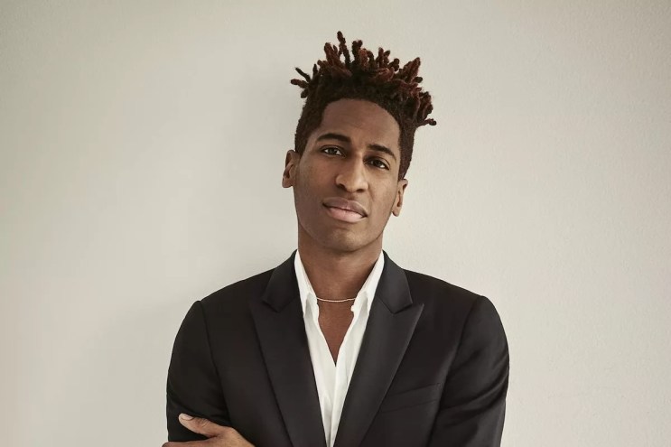 Jon Batiste in a suit standing against a beige background