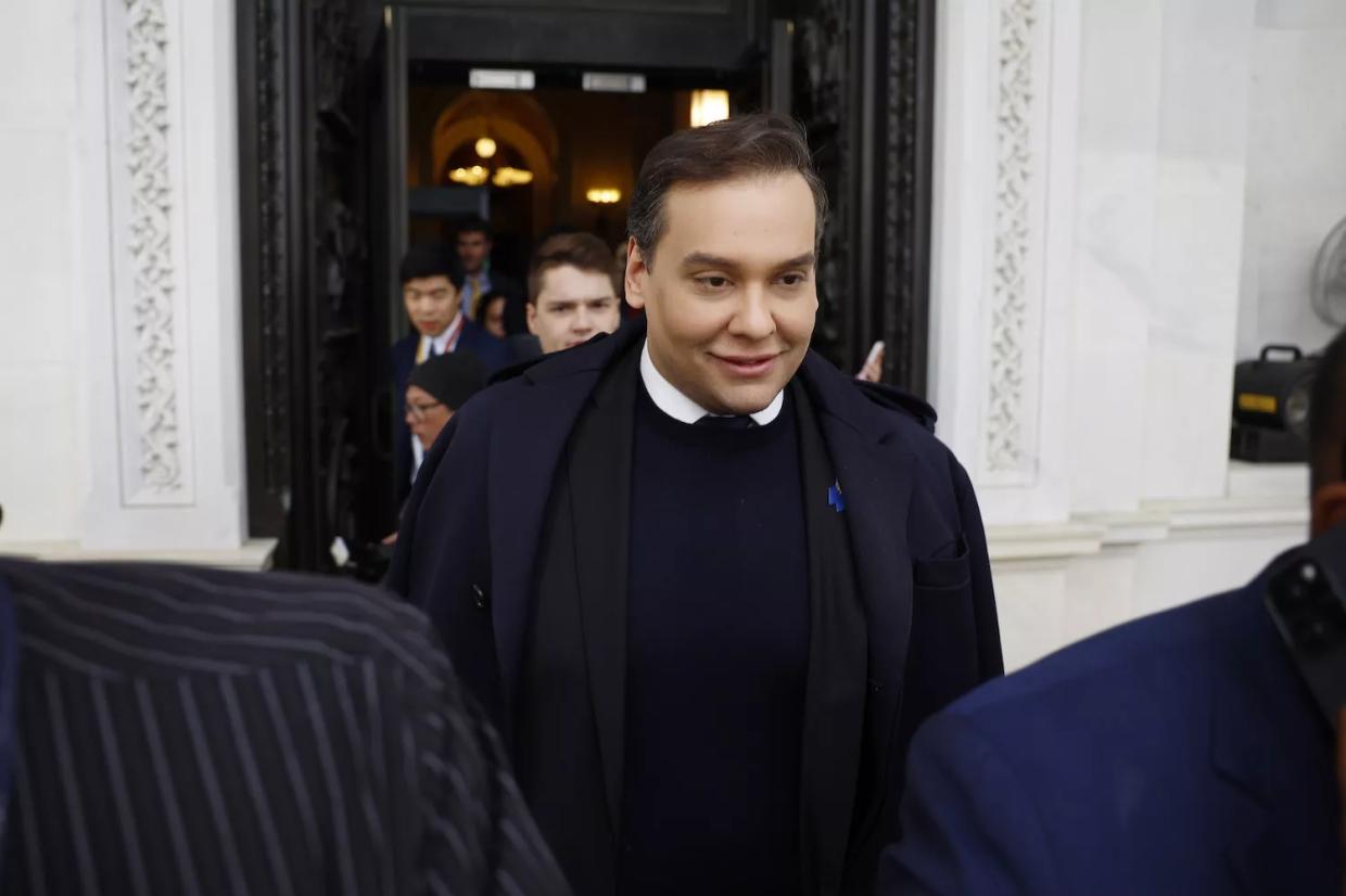 color photo of George Santos dapperly clad with a white shirt and dark tie peeking out above a navy blue sweater, smiling seemingly uncomfortably