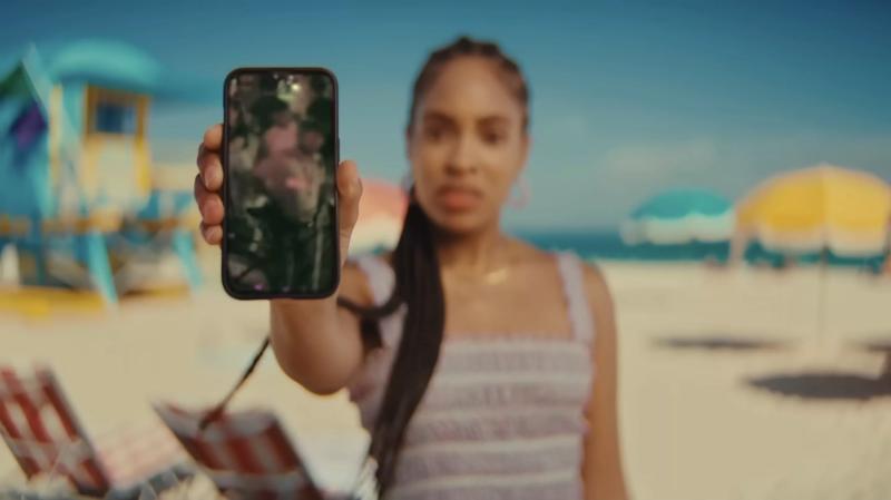 A screenshot of a Black female actor with long braids on Miami Beach holding a  smartphone up to the viewer