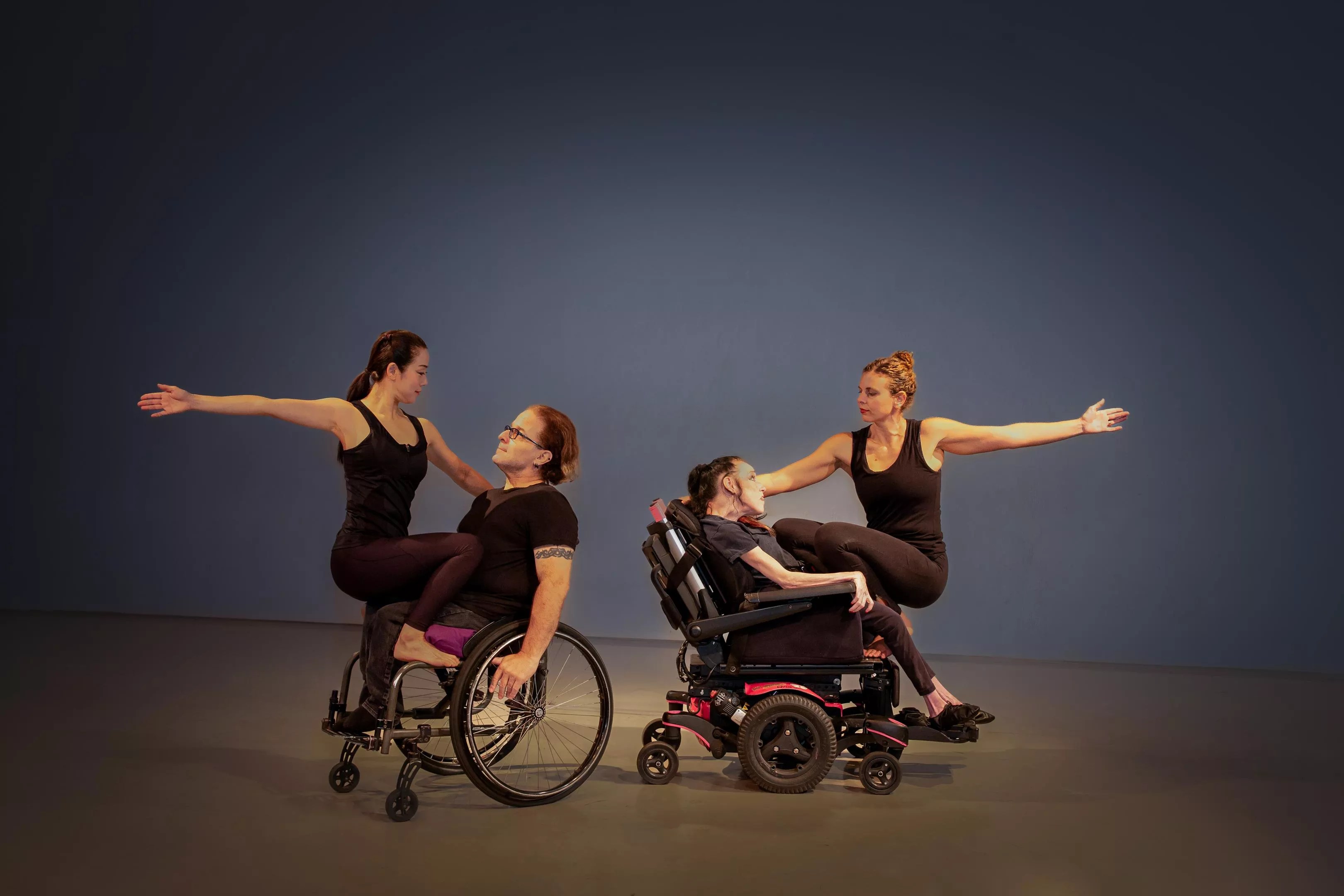 color photo of two sets of partners performing a dance on a stage; each partnership consists of one dancer who uses a wheelchair and one who does not