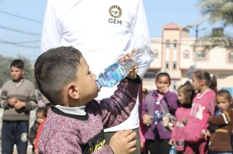 A child in Gaza drinks from a water bottle in front of a person wearing a shirt with a Global Empowerment Mission logo.