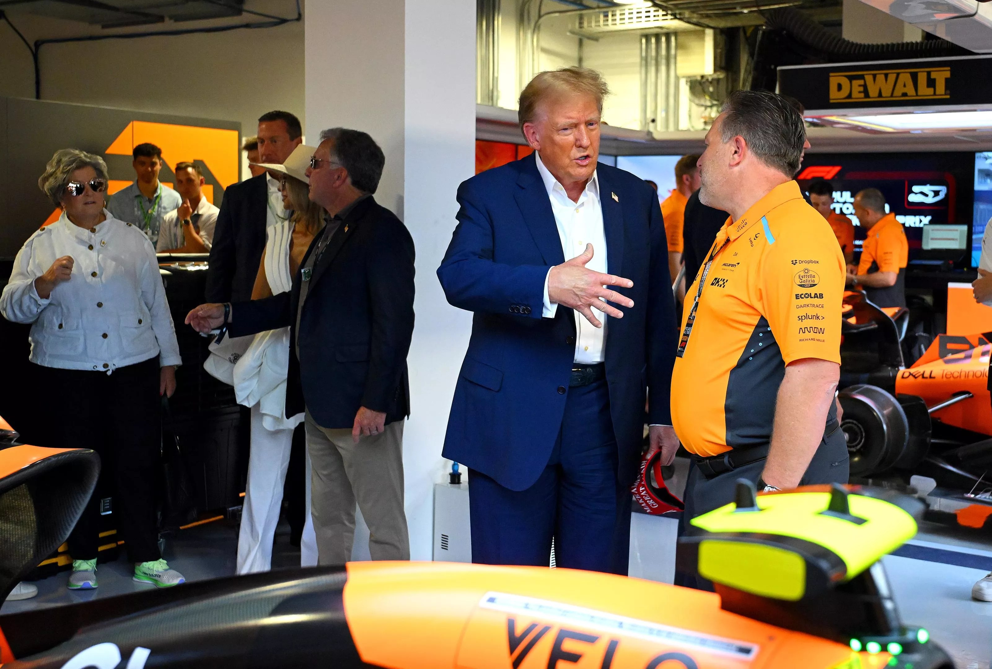 Donald Trump stands in front of a race car, talking to a McLaren executive while gesturing with his right hand