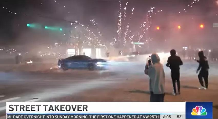 A car spins around an intersection during a street takeover as fireworks explode in the background