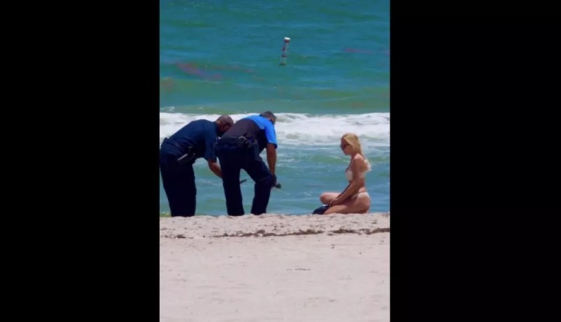 A woman in a bikini and two police officers crouch over a brick of white powder on the beach