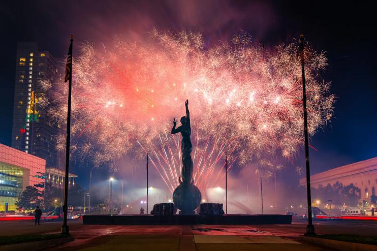 A large cluster of fireworks explode with a statue silhouette in the foreground in Cleveland