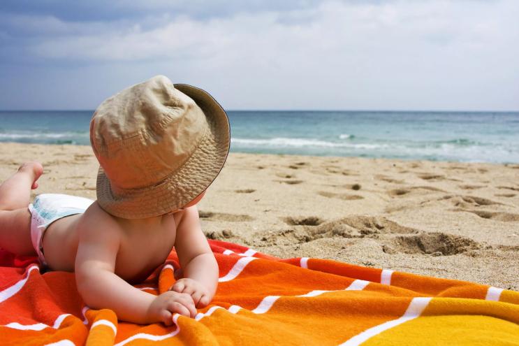 A baby on a beach towel stares out at the ocean on a sunny day