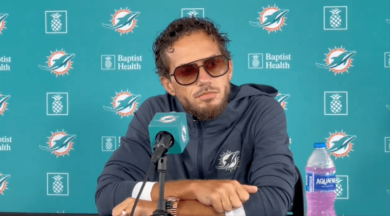 Mike McDaniel speaks at press conference with flashy shades, curly hair, and an overgrown beard.