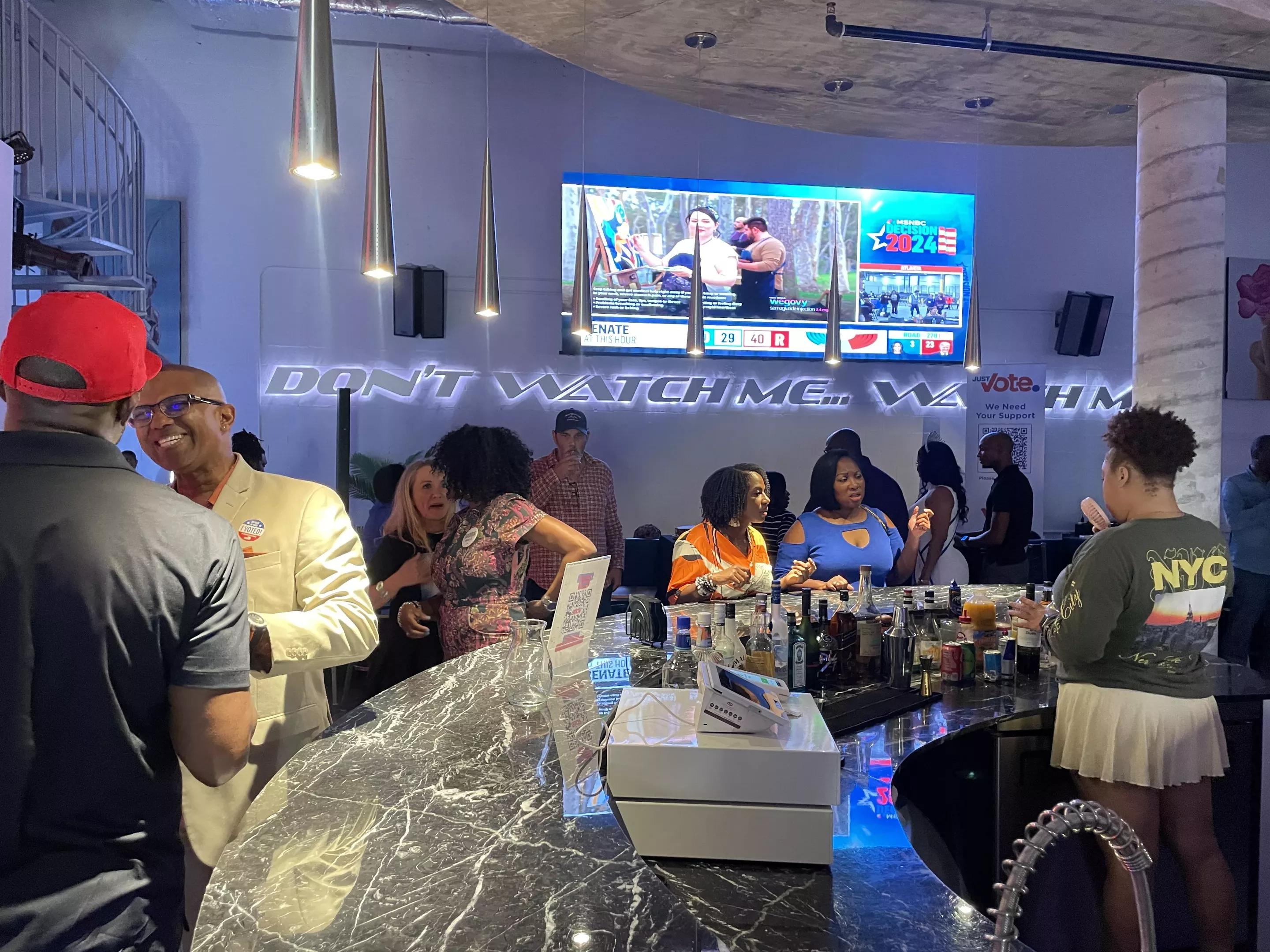 Crowd at Just Vote Period watch party gathered around the bar with a screen displaying election results in the background
