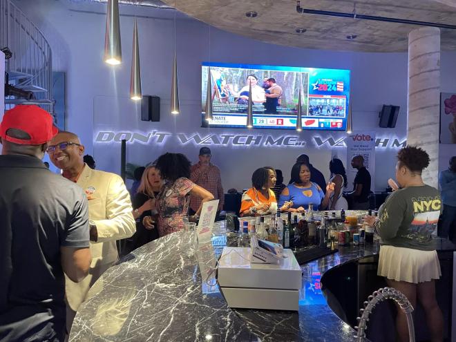 Crowd at Just Vote Period watch party gathered around the bar with a screen displaying election results in the background