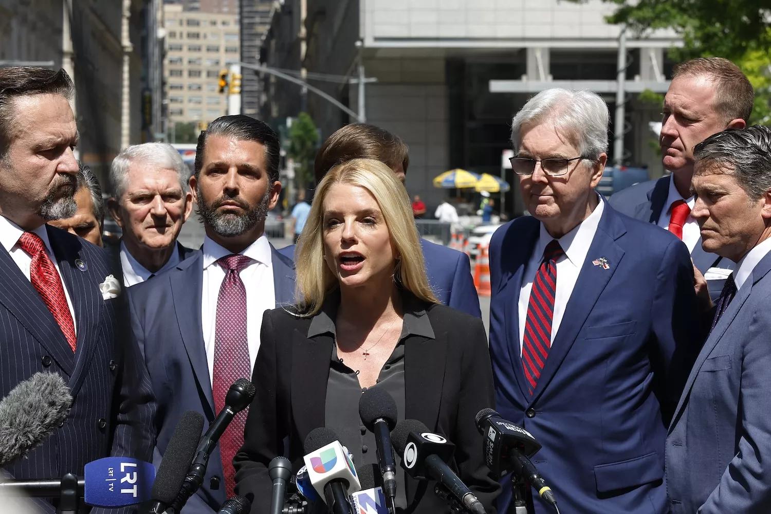 Blond woman in front of a bank of microphones with several people around her, including Donald Trump Jr.