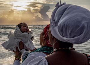 A photo by Woosler Delisfort depicting a spiritual baptism ritual with a child held at the ocean’s edge during sunrise