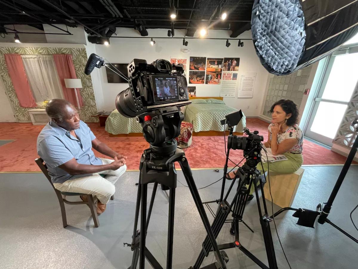 A camera stands between two a filmmaker and interviewee. A bed is part of a museum exhibition in the background.