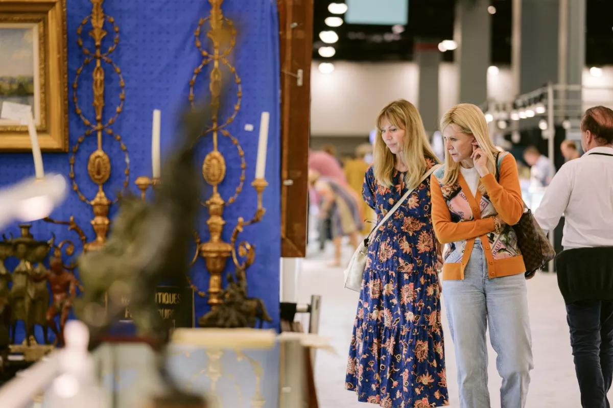 Two blonde women look at antiques including sculptures and candle holders.
