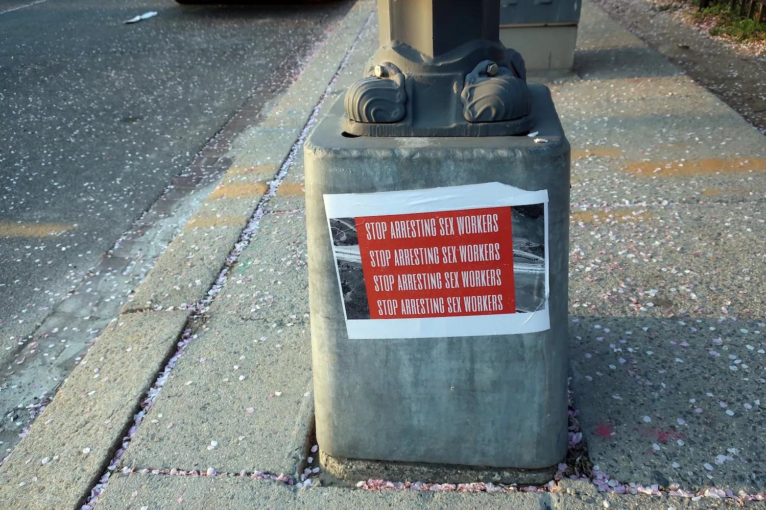 Photo of the base of a utility pole to which sign has been affixed that reads "Stop Arresting Sex Workers" in all caps, repeated four times