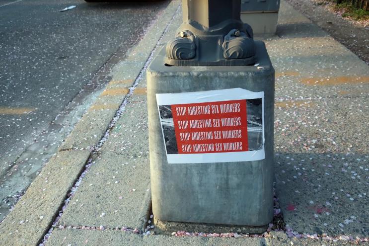 Photo of the base of a utility pole to which sign has been affixed that reads "Stop Arresting Sex Workers" in all caps, repeated four times