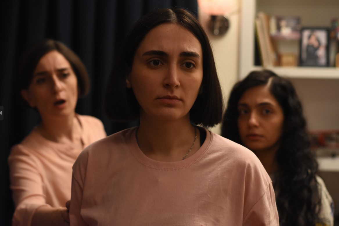 A closeup of a woman in a pink shirt. Behind her, an older woman bears an expression of concern. A young girl in the background on the right is stoic.