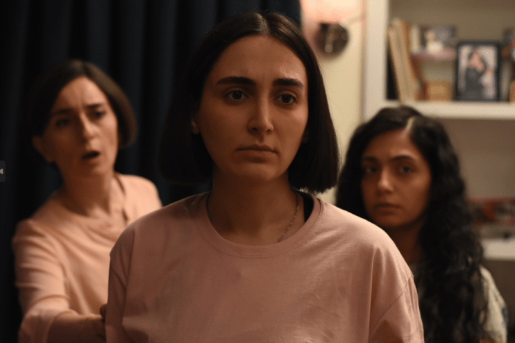 A closeup of a woman in a pink shirt. Behind her, an older woman bears an expression of concern. A young girl in the background on the right is stoic.