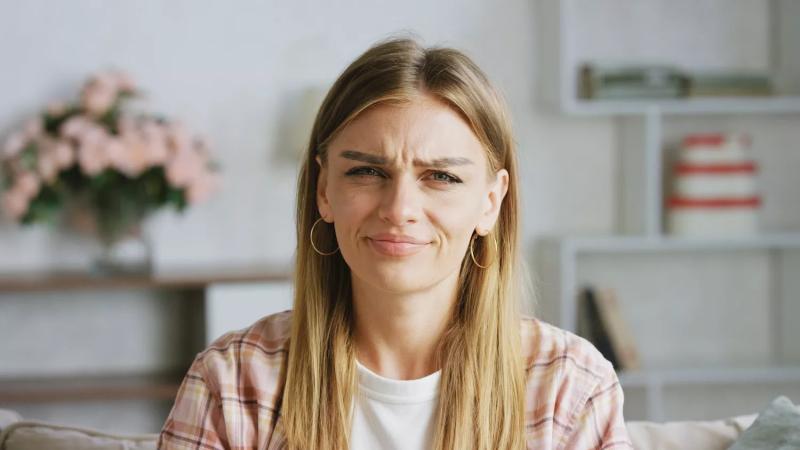Young woman wearing casual clothes frowning on a sofa in her living room, expressing clear disgust and disapproval, reflecting negative emotions and frustration
