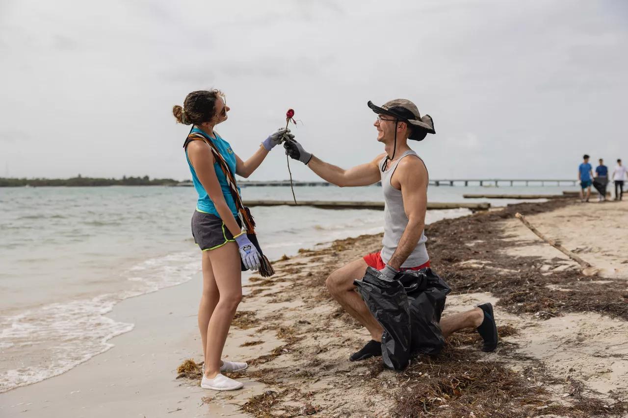 A man on one knee hands a rose to a woman on the beach. They both wear gloves and he holds a black plastic bag.