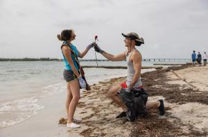 A man on one knee hands a rose to a woman on the beach. They both wear gloves and he holds a black plastic bag.