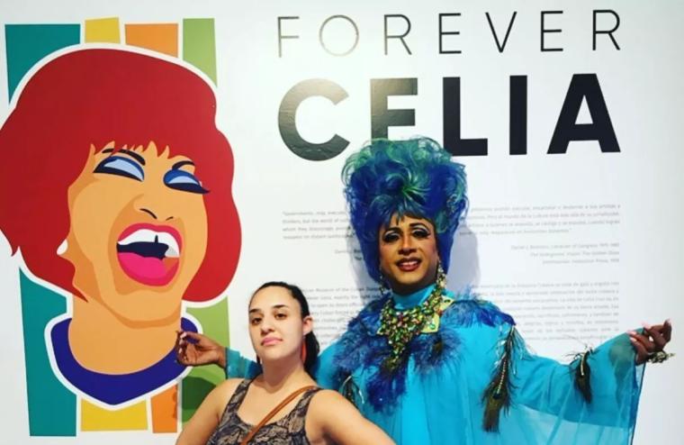 A woman and drag queen dressed like Celia Cruz pose in front of a mural of Celia Cruz. The mural reads, "Forever Celia."