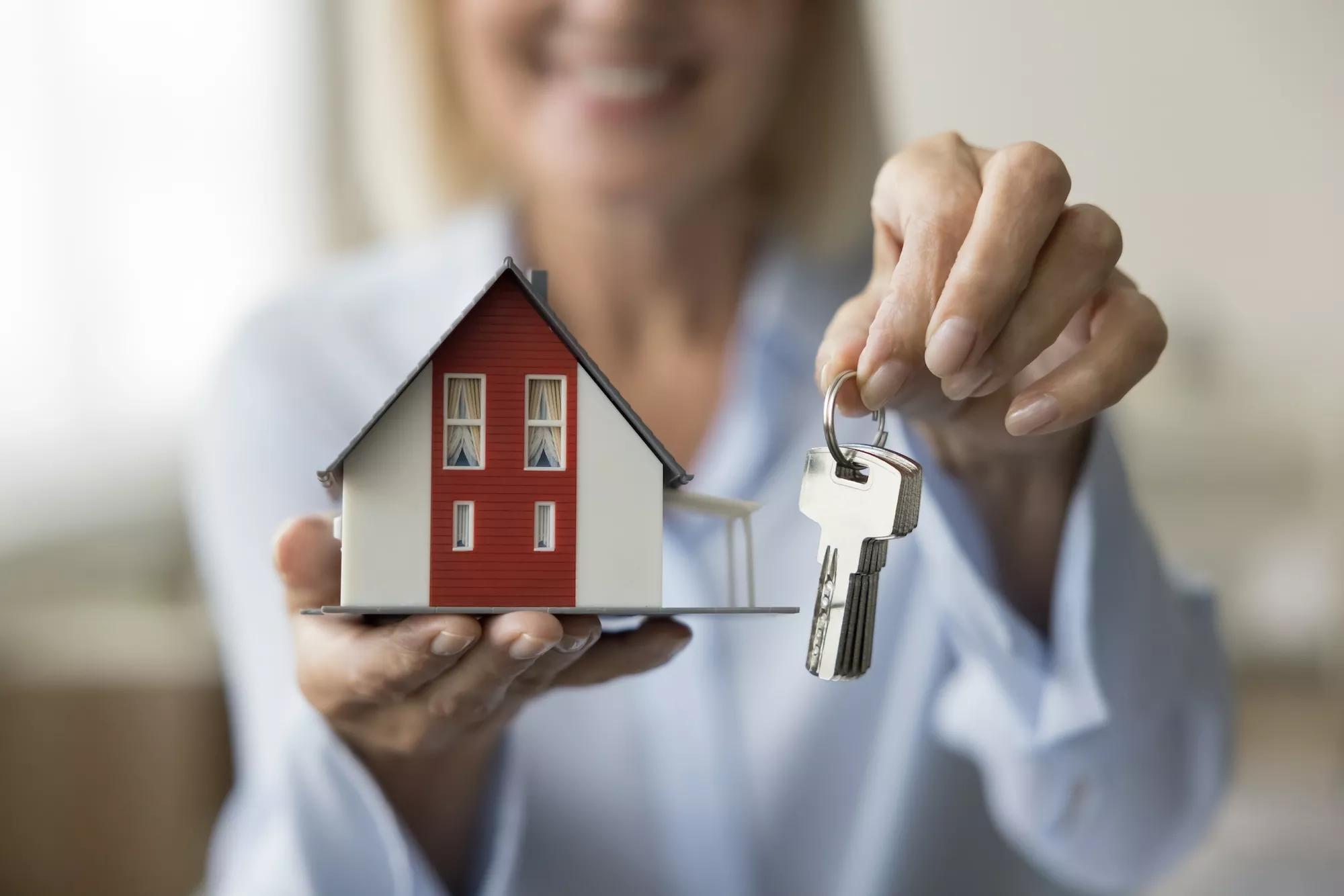 Close up of realtor holding keys and small model of family house in hands, offering apartment for rent, selling, buying.