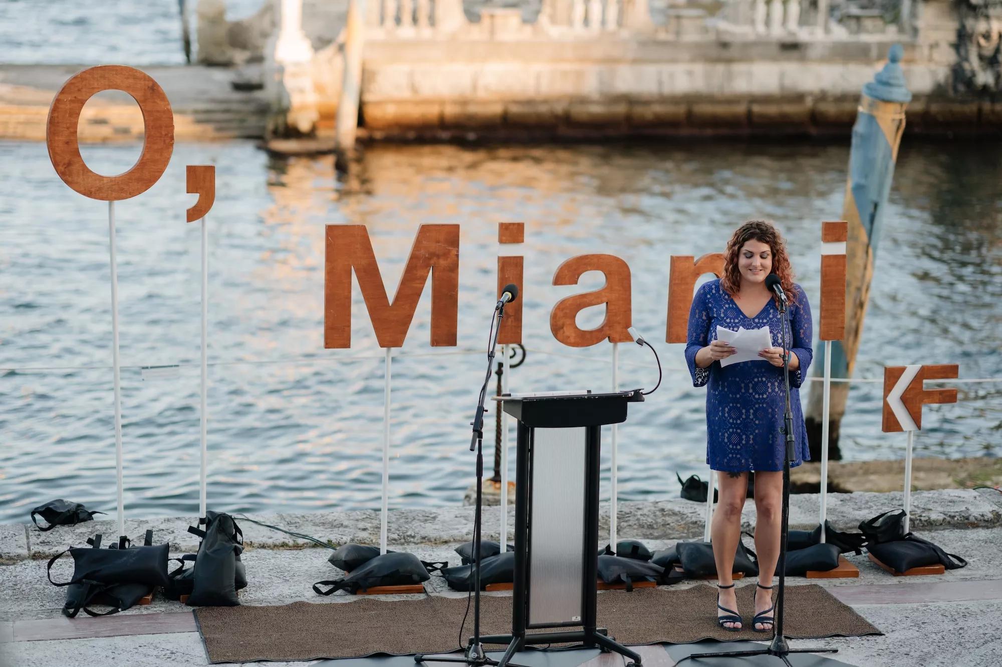 a person reads a poem in front of the water and a backdrop that reads, "O, Miami"