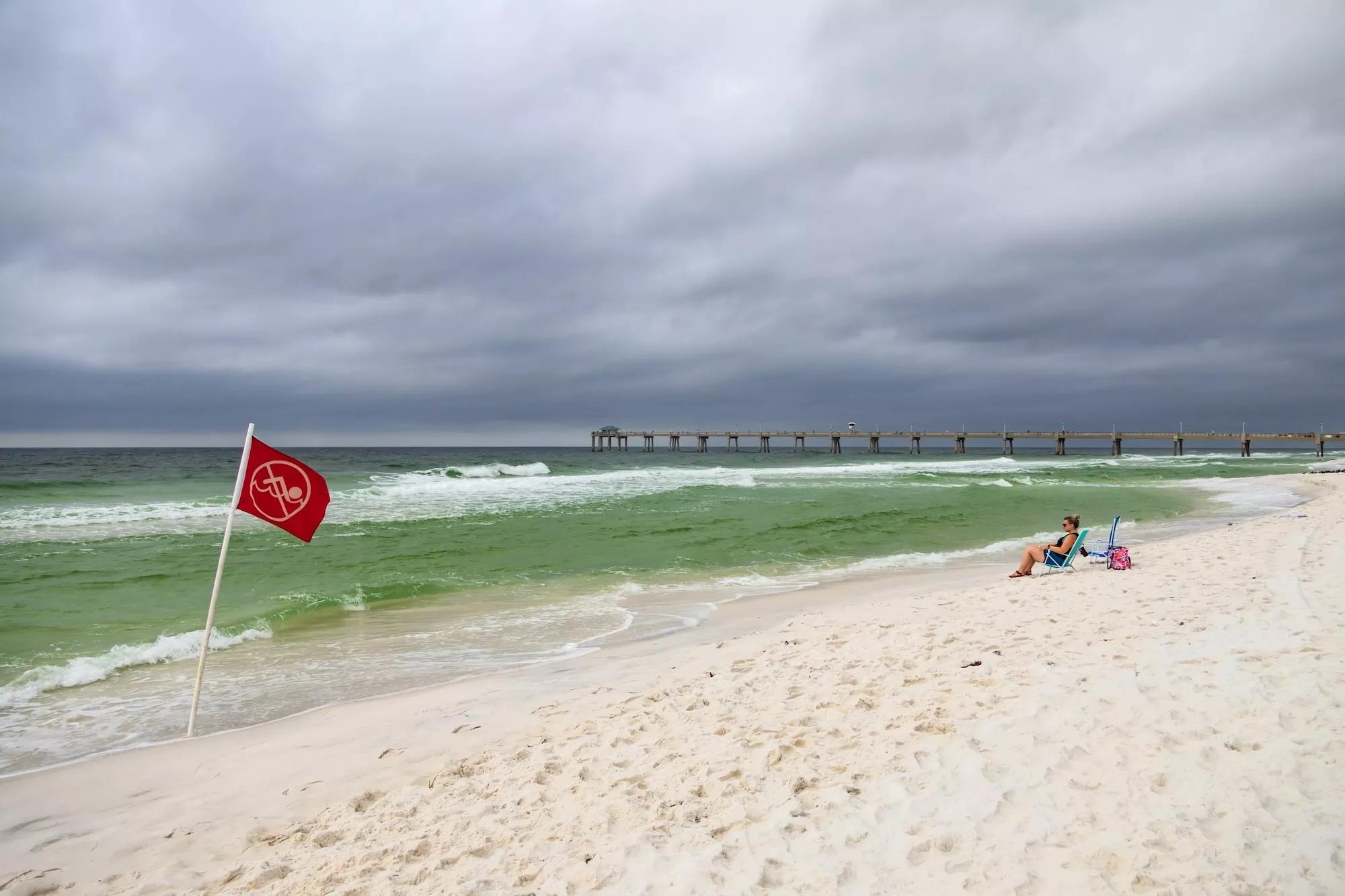 a woman sitting on a beach with stormy weather rolling in; a red flag in the foreground