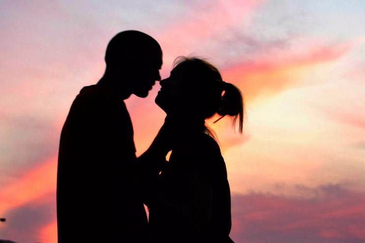 A silhouetted couple leans in for a kiss with pink golden-hour skies behind them