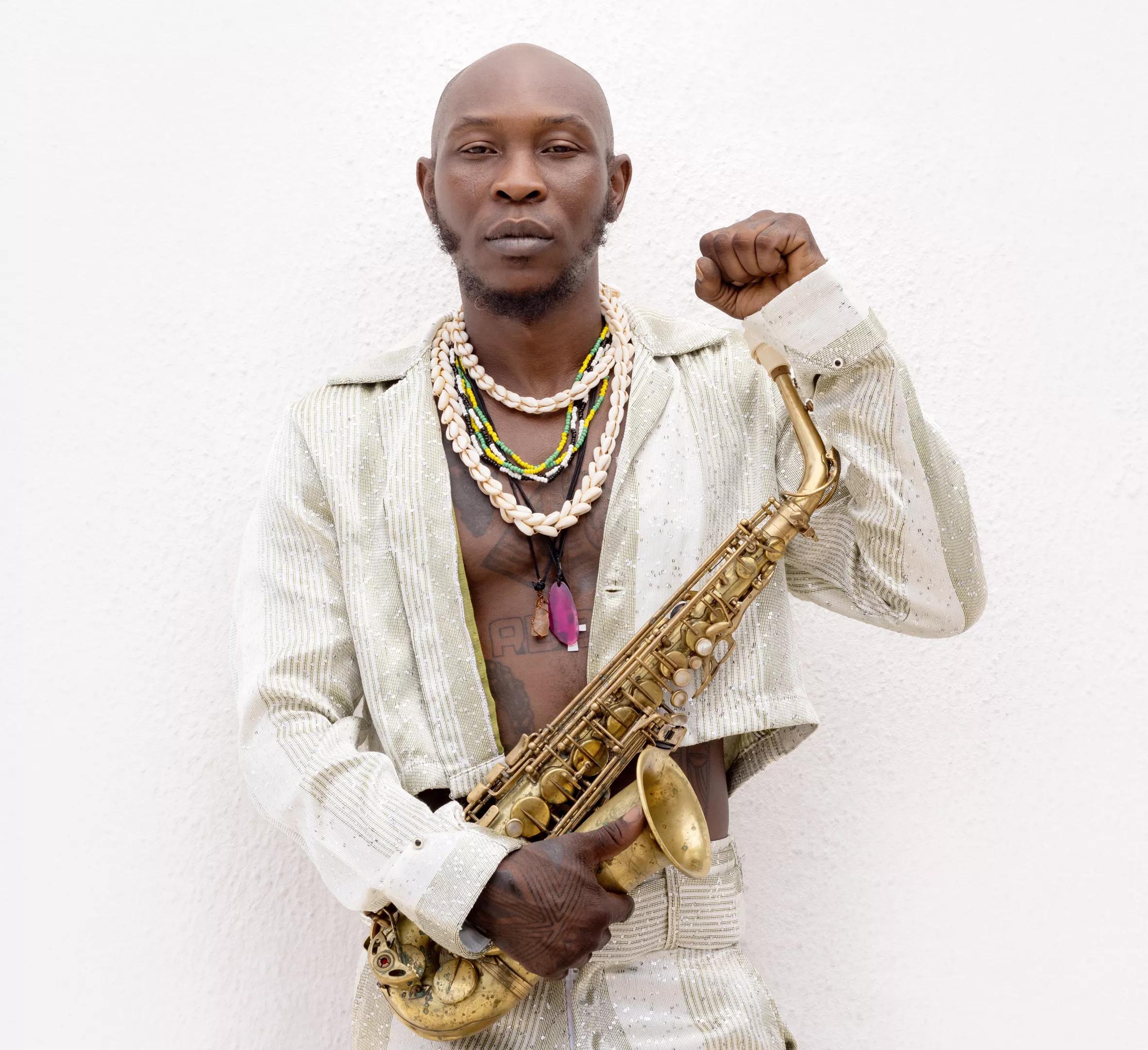Portrait of a bald, Black man dressed in white with his shirt open, wearing shell necklaces, holding a saxophone, and making a fist