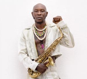 Portrait of a bald, Black man dressed in white with his shirt open, wearing shell necklaces, holding a saxophone, and making a fist