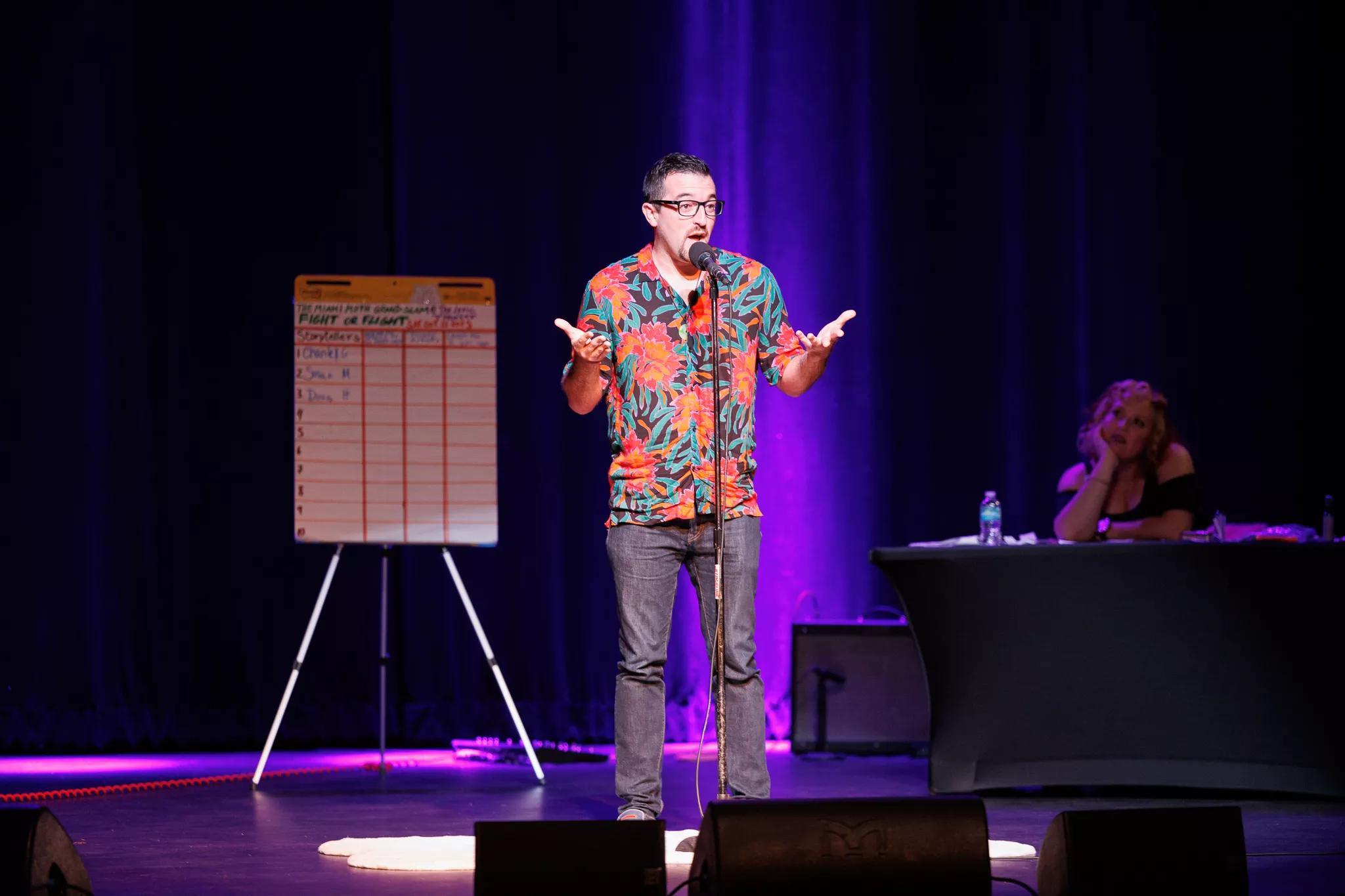 photo of a man speaking on stage with a scoreboard behind him on a whiteboard