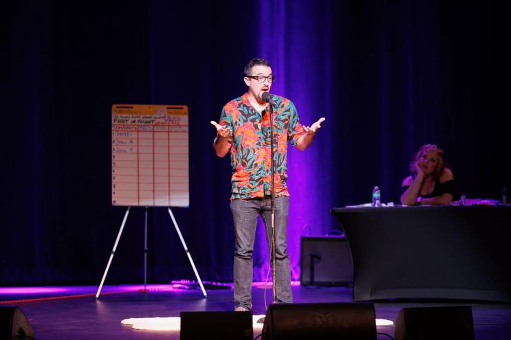 photo of a man speaking on stage with a scoreboard behind him on a whiteboard