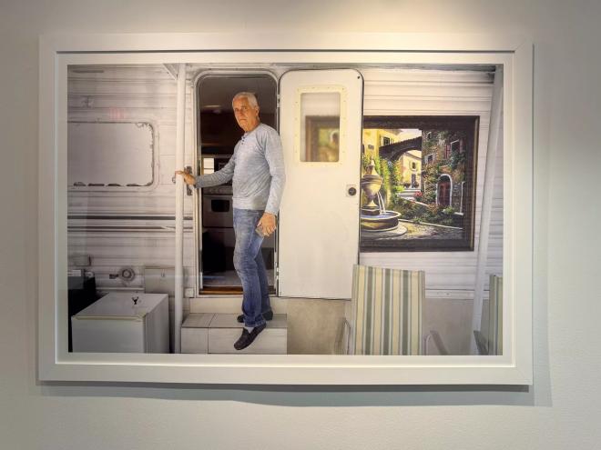 A framed photo is on display in a gallery. In the image, a man stands at the entrance of his mobile home. A painting with a fountain and greenery hangs next to the door